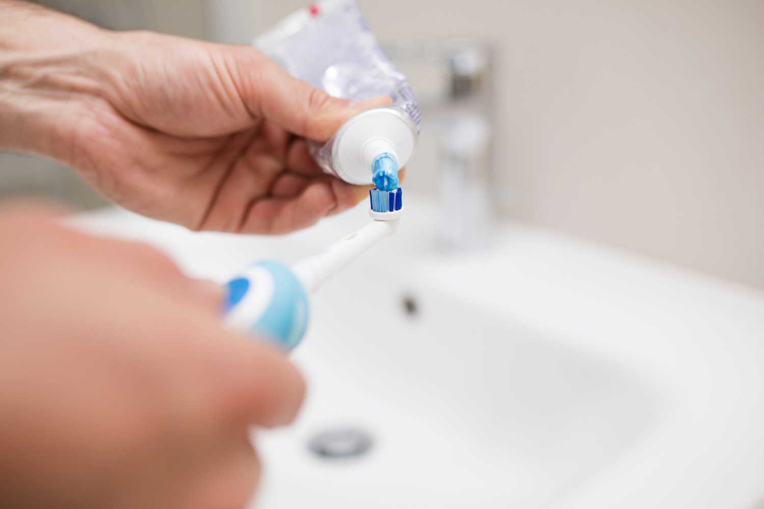 Person applying toothpaste to an electric toothbrush over the sink